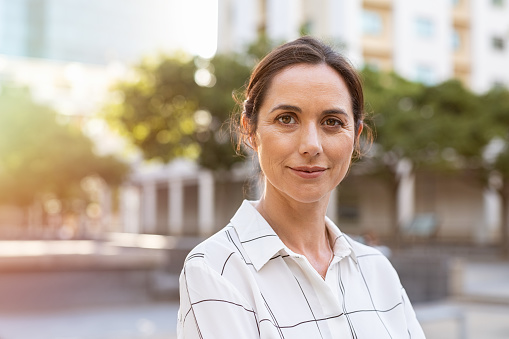 Portrait of happy mature businesswoman looking at camera. Successful proud woman in city street at sunset. Satisfied latin business woman in formal clothing smiling outdoors.